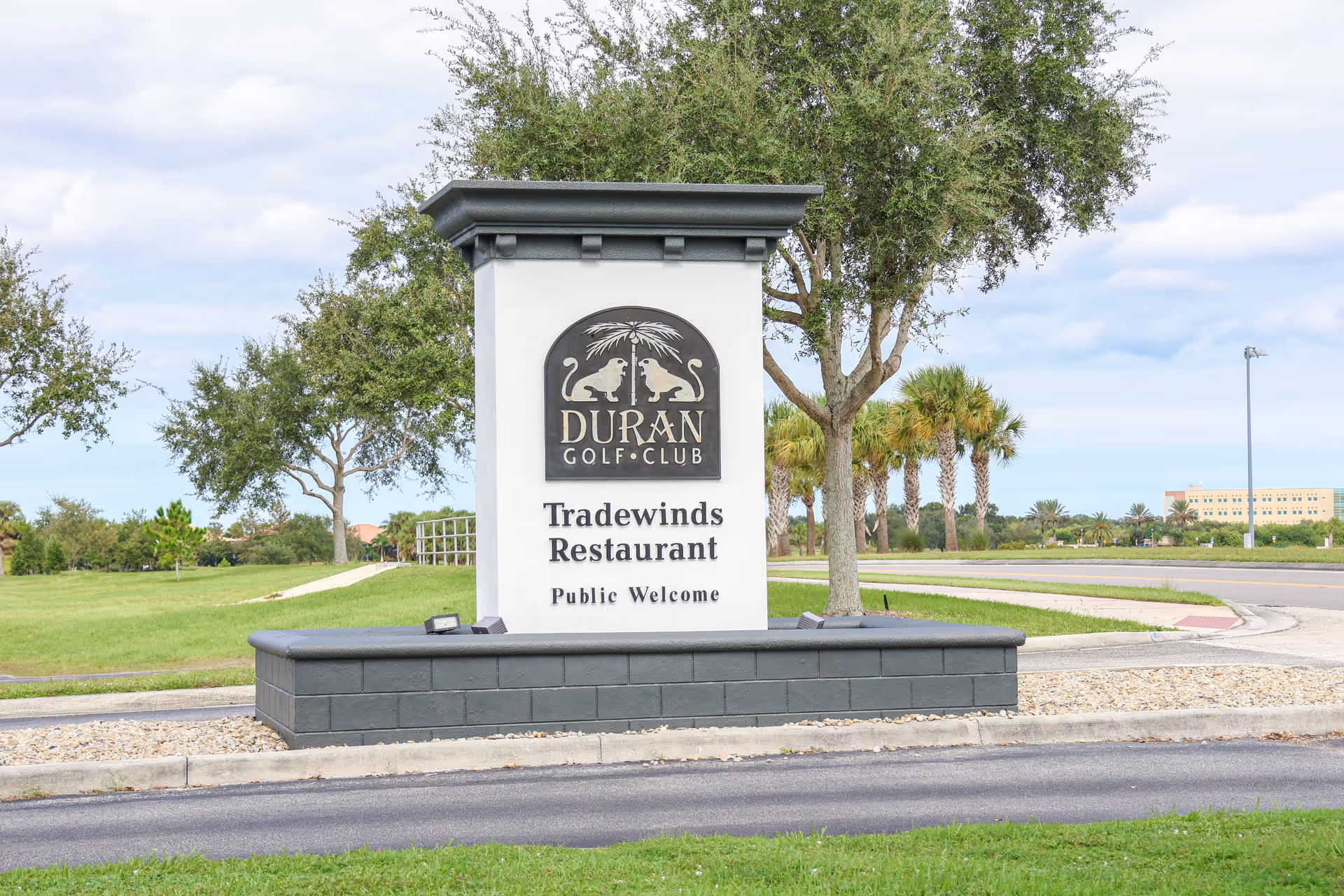 Stone entrance sign reading 'Duran Golf Club Tradewinds Restaurant Public Welcome' with trees, lawn and a road in the background.