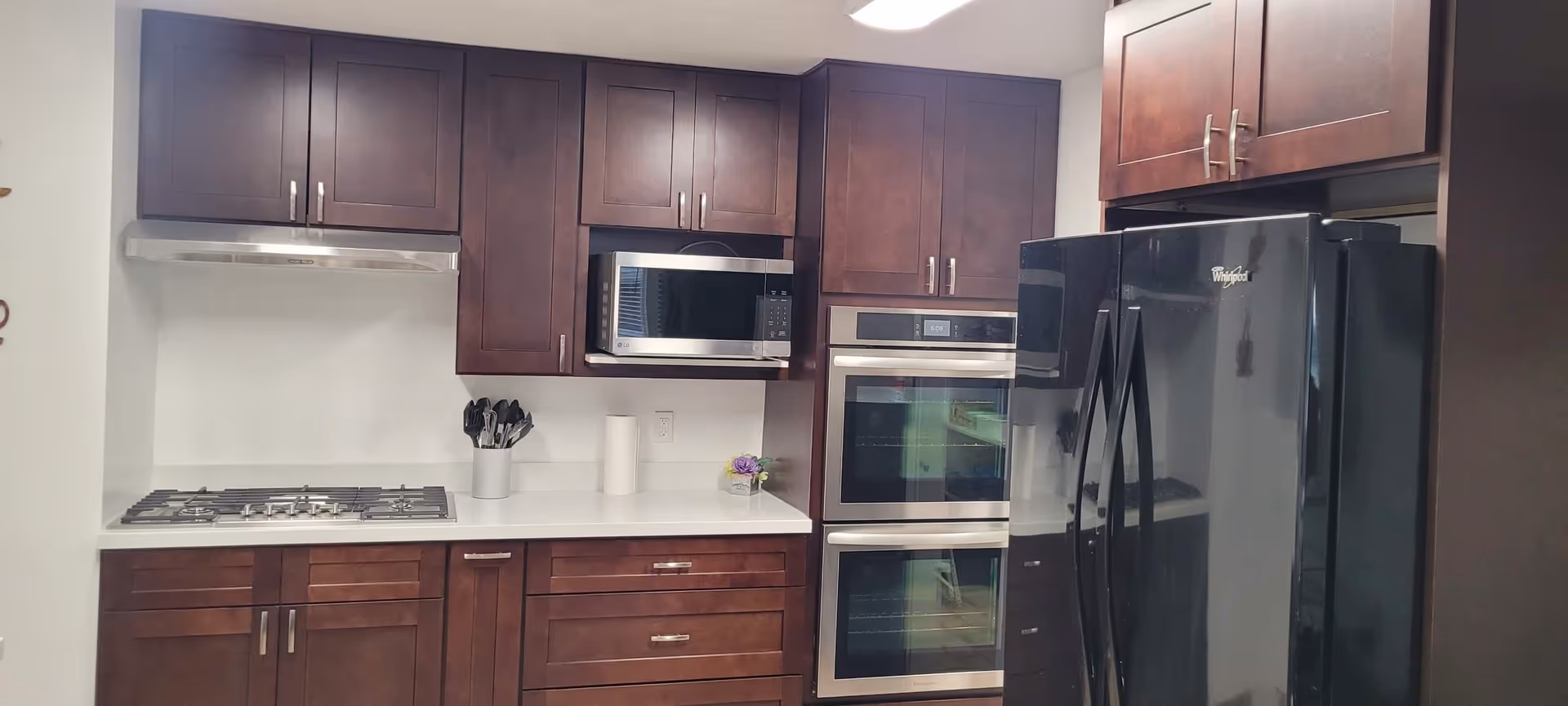 Modern kitchen with dark wood cabinets, a stainless steel gas stove with a hood, a microwave, double wall ovens, and a black Whirlpool refrigerator.