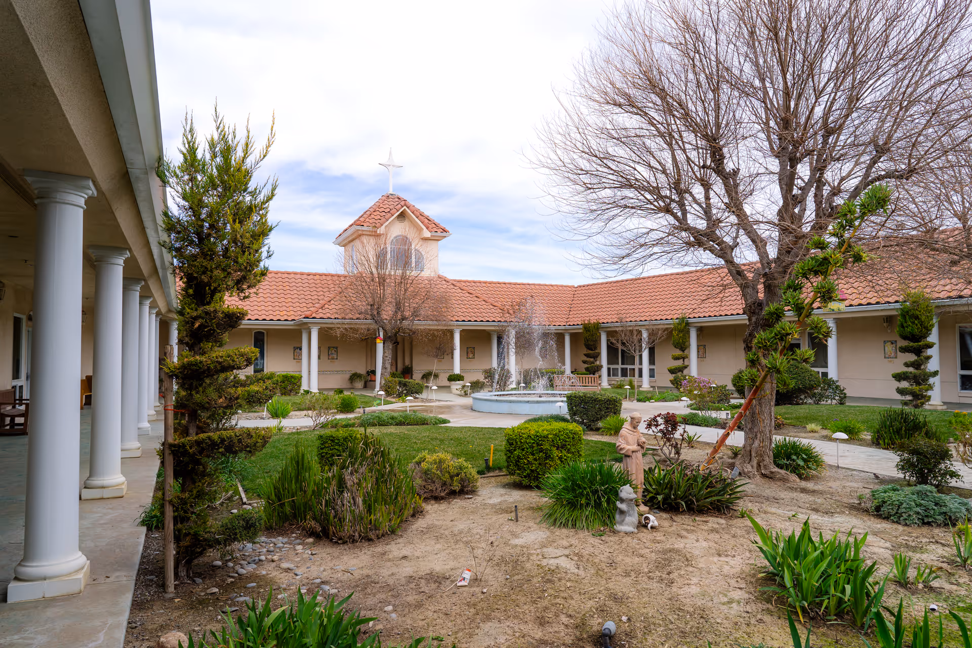 Outdoor courtyard of New Bethany Residential Care featuring a central fountain, manicured bushes, trees with no leaves, a statue, and a building with a tiled roof and white columns surrounding the courtyard.