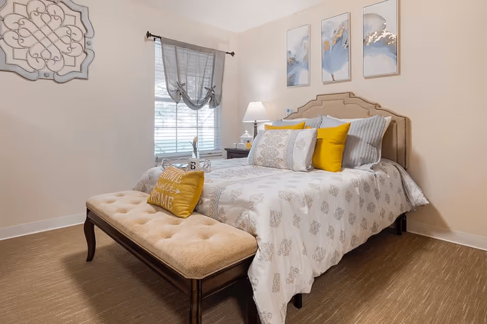 A neatly made bed with a beige upholstered headboard, white and patterned bedding, and yellow accent pillows in a cozy bedroom. A cushioned bench with a yellow pillow sits at the foot of the bed. There is a window with sheer curtains, a nightstand with a lamp, and three framed artworks on the wall above the bed.