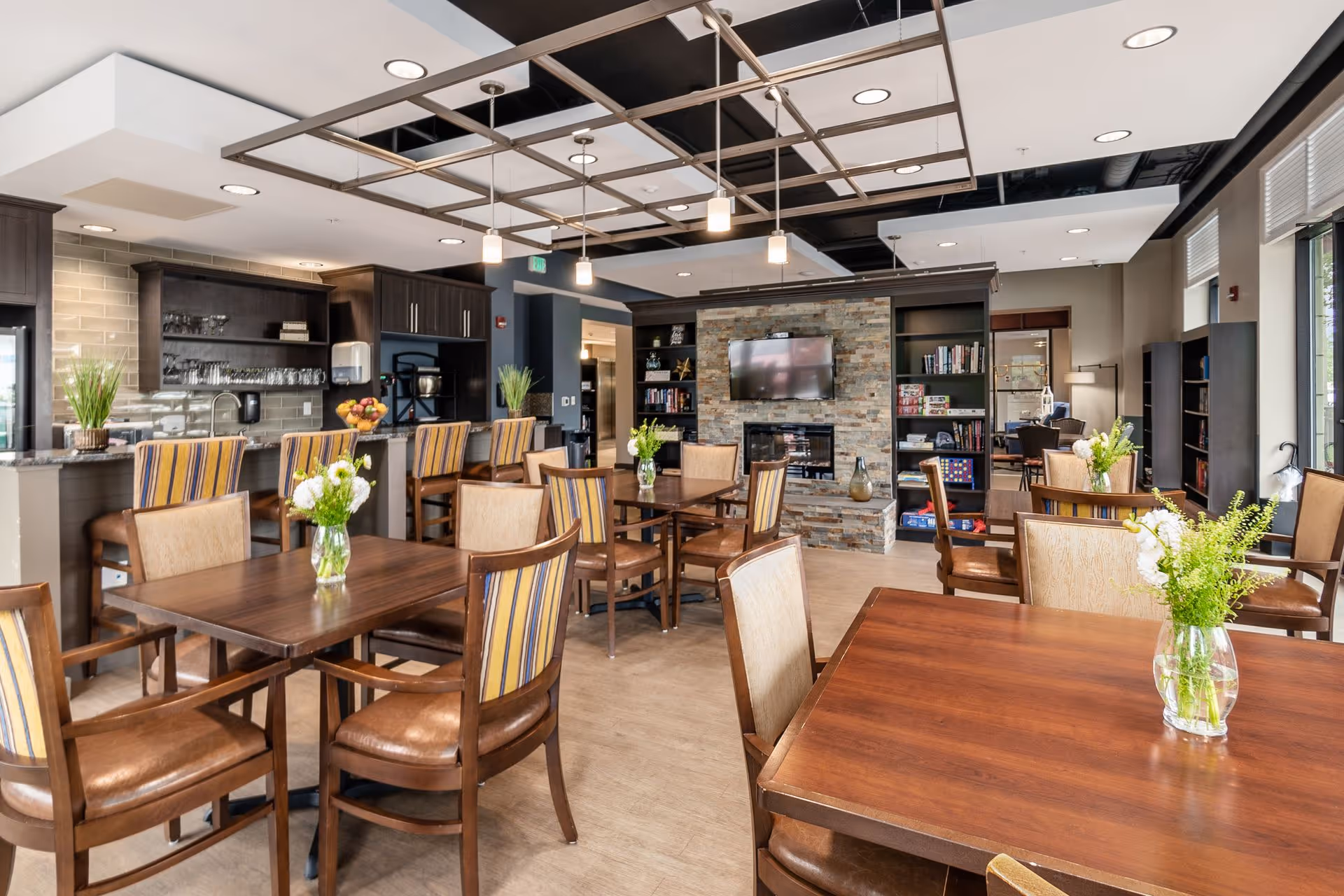 Spacious communal dining and lounge area with wooden tables and striped-back chairs, a bar, and a stone fireplace with a mounted TV.