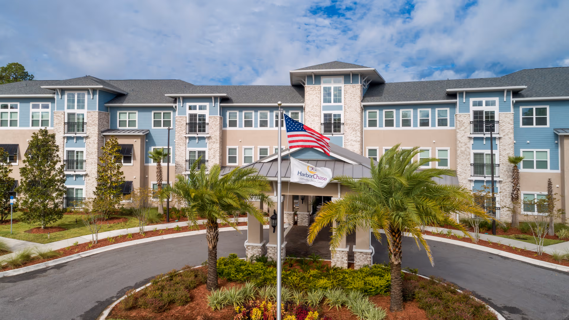 Front exterior view of HarborChase of Mandarin, a multi-story senior living facility with a circular driveway, landscaped greenery, palm trees, and an American flag flying on a flagpole in front of the entrance.