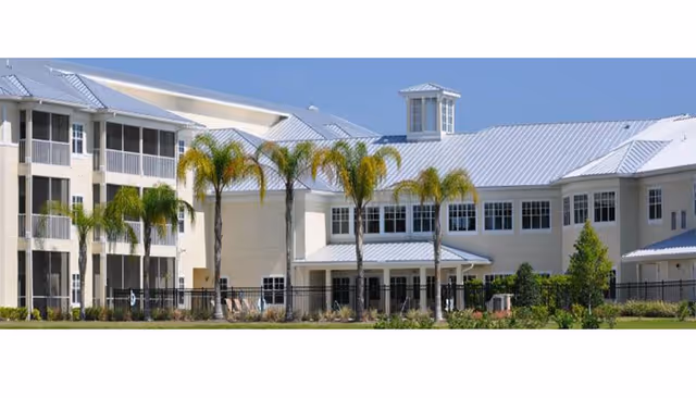 Exterior front of a multi-story senior living facility with metal roofs, balconies, palm trees, and landscaped grounds.