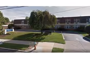 Exterior view of Liberty Center for Rehabilitation and Nursing, a two-story building with a curved driveway and a grassy area with a tree and a bench in front. There is a sidewalk and a fire hydrant near the street.