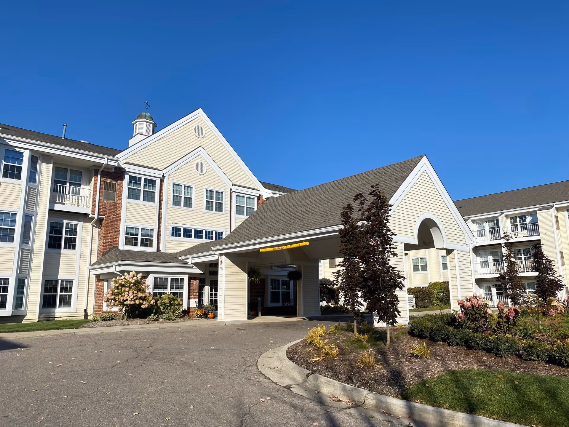 Exterior view of a senior living facility building with beige siding and brick accents under a clear blue sky. The building has multiple windows and a covered entrance with a driveway. There are landscaped areas with small trees and flowering plants near the entrance.