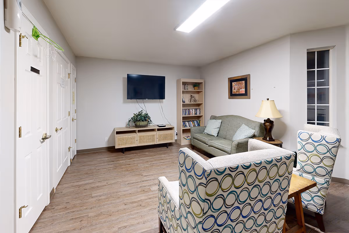 A cozy living room with a green sofa, two patterned armchairs, a wooden side table with a lamp, a wall-mounted flat screen TV, a wooden media console with storage baskets, and a bookshelf filled with books and decorative items. The room has light-colored walls, wood flooring, and a small window with multiple panes.