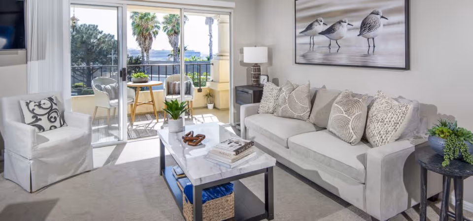 A bright living room with a light gray sofa adorned with multiple patterned cushions, a white armchair with a decorative pillow, and a marble-top coffee table holding books and decor items. Behind the sofa is a large framed picture of three shorebirds. Sliding glass doors open to a balcony with two wicker chairs and a small round table, with palm trees visible outside.
