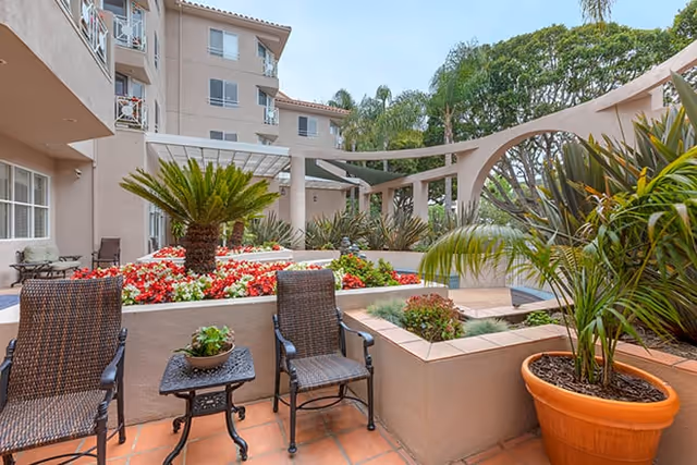 Outdoor patio area at Brookdale Santa Monica Gardens featuring two wicker chairs with a small table between them, surrounded by potted plants and flower beds. The background shows a multi-story building with balconies and a pergola structure over part of the patio.
