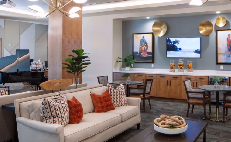A modern living room area with a beige sofa adorned with patterned and solid red pillows. Behind the sofa, there is a black grand piano and a wooden chair. The room features a wall with a mounted TV flanked by two framed artworks and decorative gold wall accents. Below the TV is a countertop with cabinets and glass containers filled with oranges. There are also round tables with chairs arranged in the space, and a coffee table in the foreground with a decorative bowl containing intertwined wooden pieces.