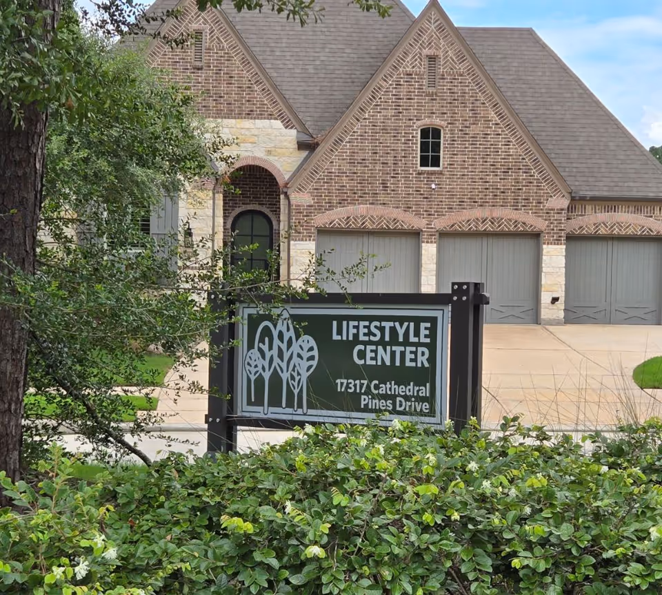 A brick and stone building with a three-car garage behind a sign that reads 'Lifestyle Center 17317 Cathedral Pines Drive,' surrounded by green bushes and trees.