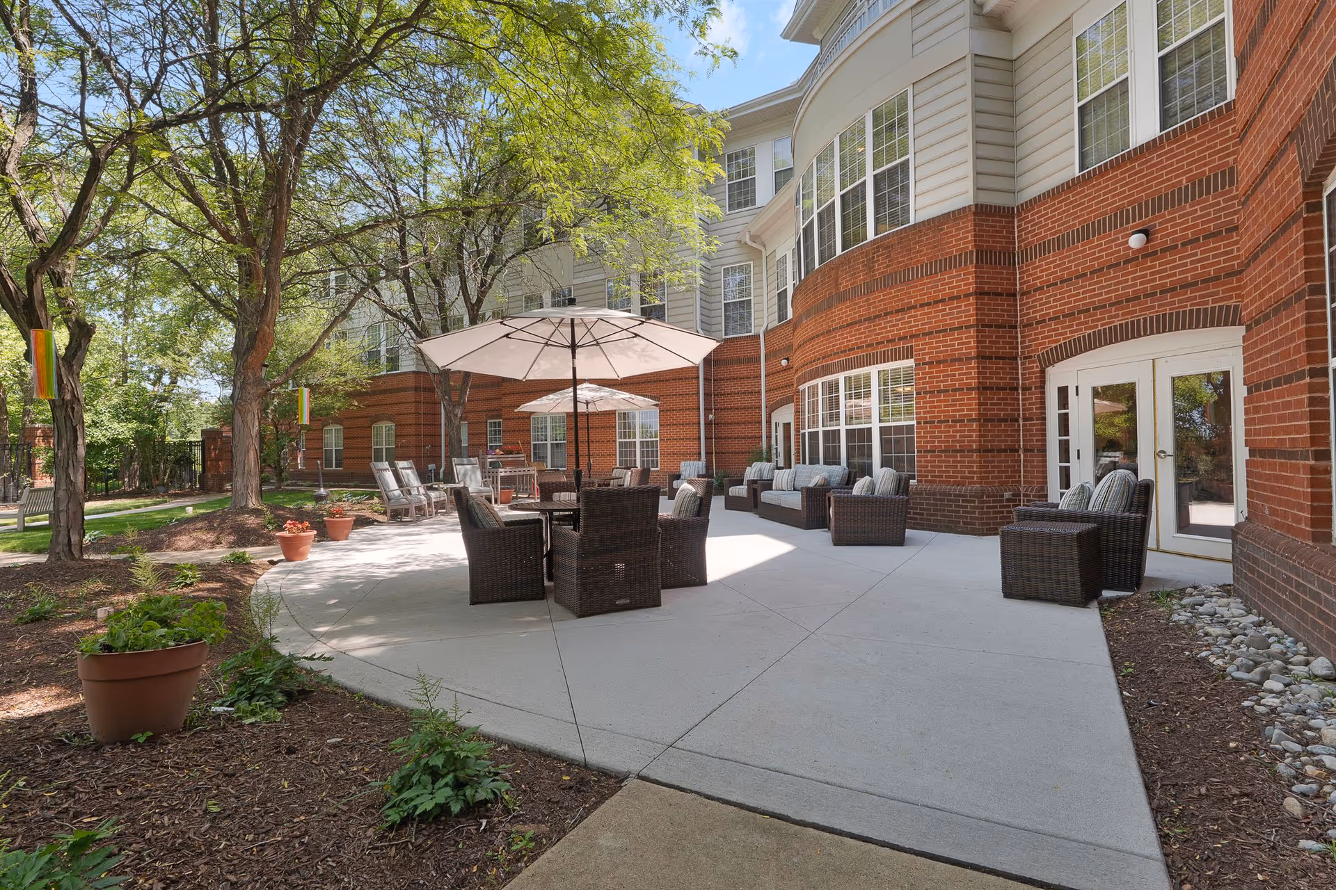 Outdoor patio area at Sunrise at Reston Town Center with wicker chairs and tables under large umbrellas, surrounded by trees and plants, adjacent to a red brick and beige siding building with large windows.