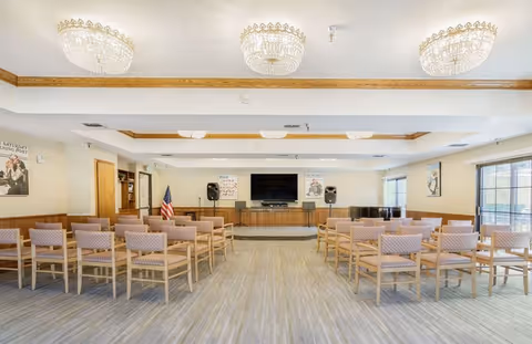 Bright multipurpose activity room with rows of chairs facing a stage area with a wall-mounted TV, speakers, and a piano.
