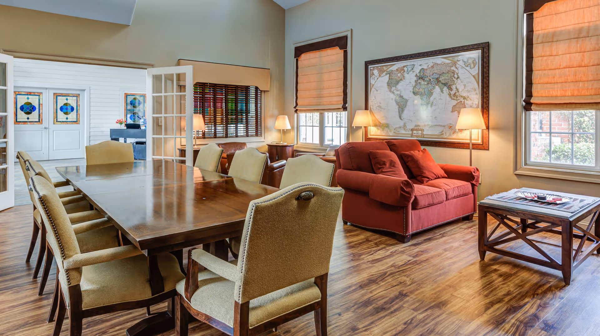 A cozy interior room featuring a large wooden dining table surrounded by eight upholstered chairs. To the right, there is a red loveseat with two matching pillows, positioned beneath a large framed world map. Two windows with orange Roman shades allow natural light to fill the room. A wooden coffee table with magazines sits in front of the loveseat. The room has wooden flooring and is softly lit by several table lamps. In the background, French doors open to another room with stained glass windows.