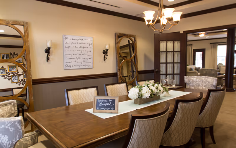 A warmly decorated dining room with a wooden table surrounded by upholstered chairs. The table has a white runner and a floral centerpiece. On the wall are two decorative mirrors and a framed script artwork. A chandelier hangs from the ceiling, and through an open doorway, a living area with armchairs and large windows is visible.