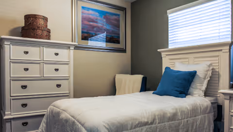 Small bedroom with a single bed made with white bedding, a white dresser, framed artwork, and a window with blinds.