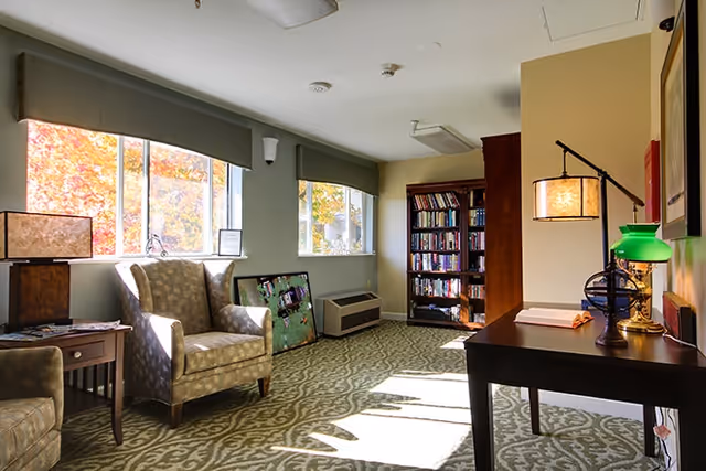 A cozy interior room with two upholstered armchairs, a wooden side table with a lamp, a bookshelf filled with books, and a desk with two lamps. Sunlight streams through two large windows, casting shadows on the patterned carpeted floor.