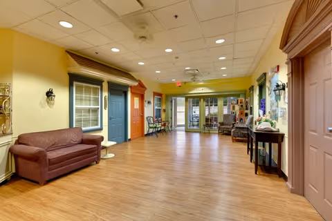 A spacious, well-lit interior hallway in a senior living facility with wood flooring and a drop ceiling with recessed lighting. The walls are painted yellow and decorated with several doors painted in different colors resembling house fronts. There is a maroon couch on the left side and a small table with flowers on the right. At the far end, glass doors lead to another room labeled 'OFFICE/RECEPTION'.