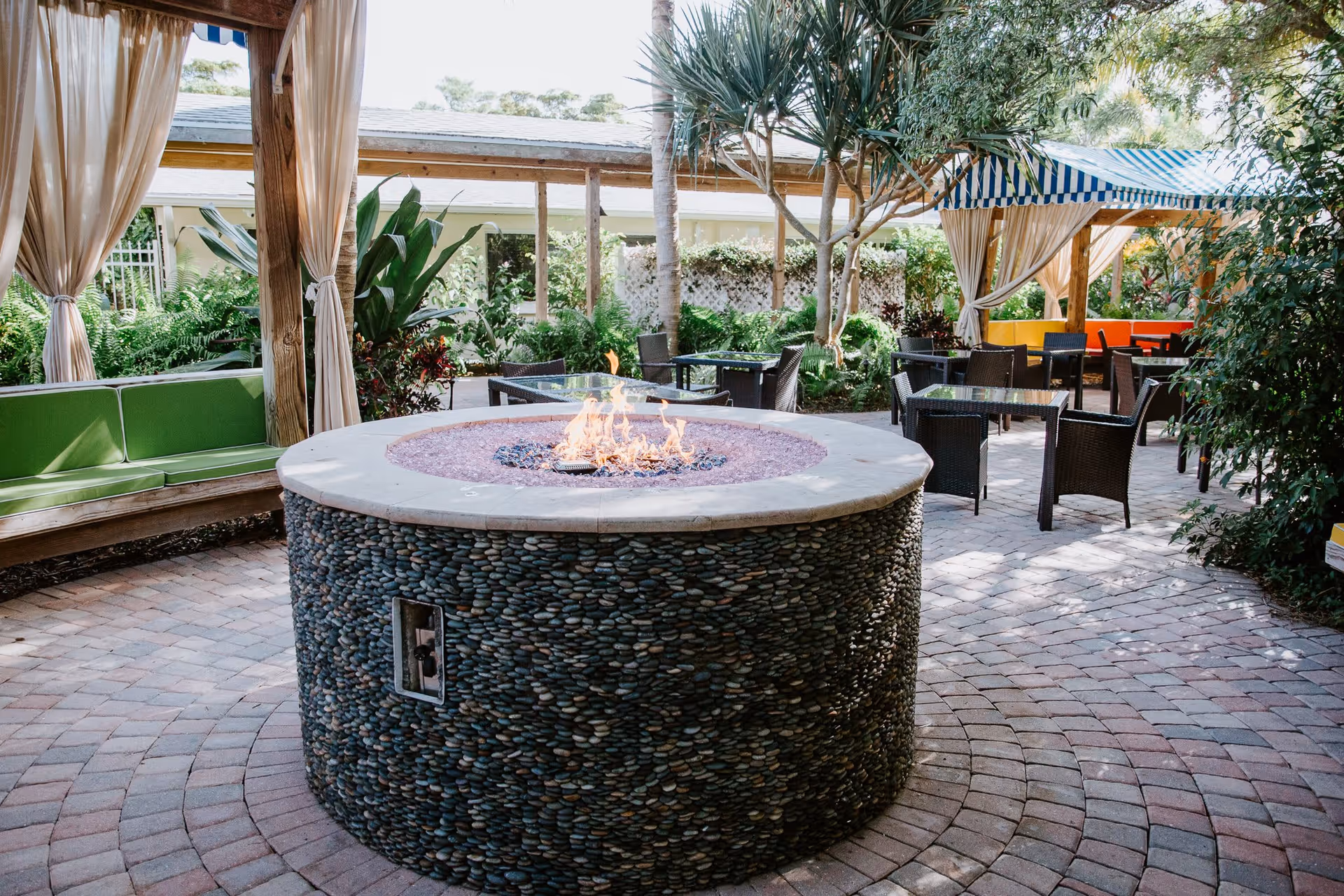 Outdoor seating area with a round stone fire pit in the center, surrounded by wicker chairs and a wooden pergola with curtains. There are green plants and trees around, and a striped blue and white canopy covering some seating in the background.