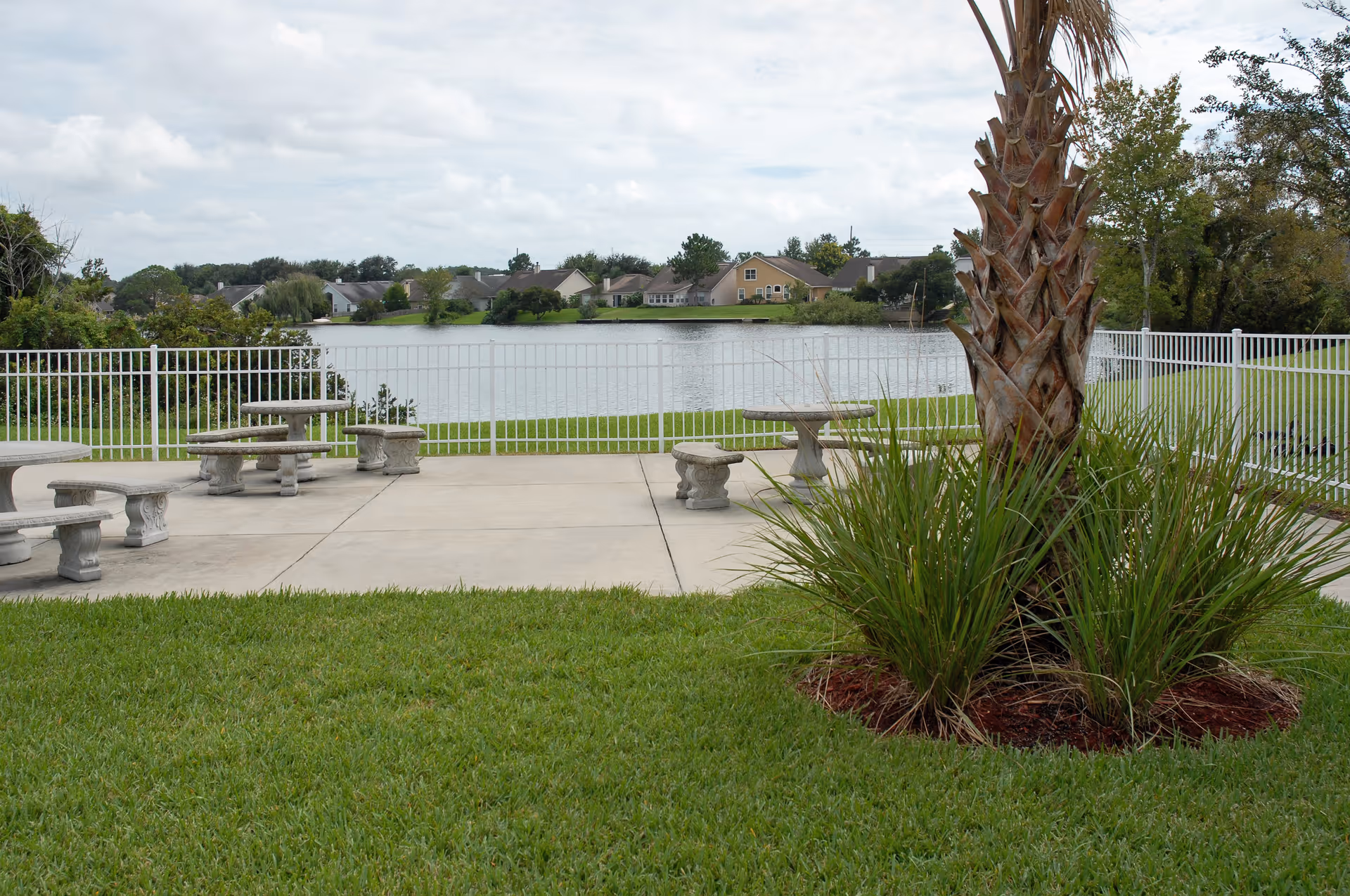Concrete patio with stone benches and tables by a white fence overlooking a lake, with grass, a palm tree, and houses across the water.