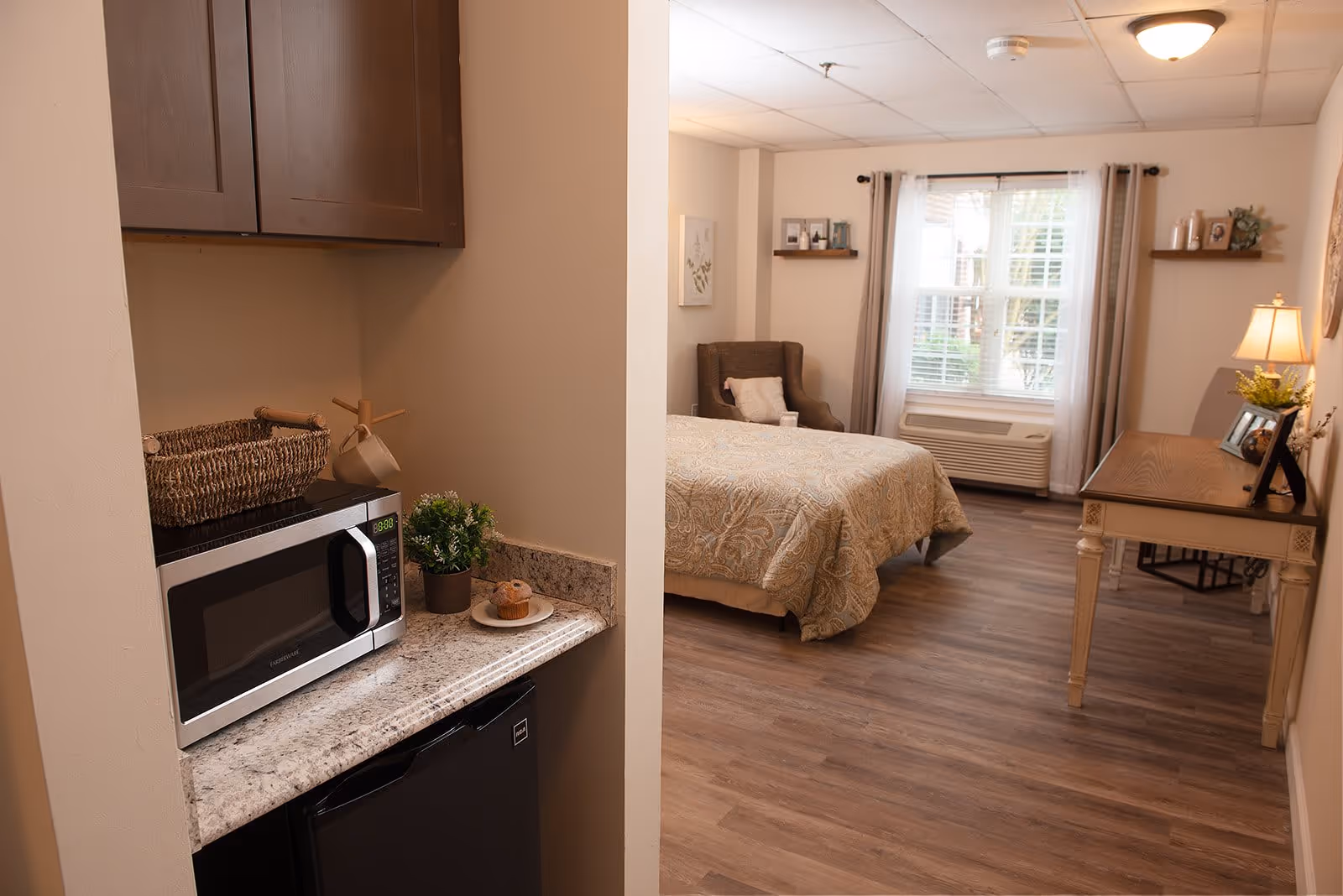 A senior living facility room featuring a small kitchenette area with a microwave, mini fridge, and countertop decorated with a basket, plant, and plate with muffins. The room also includes a bed with a patterned bedspread, an armchair, a window with curtains, a desk with a lamp and framed photos, and wooden flooring.