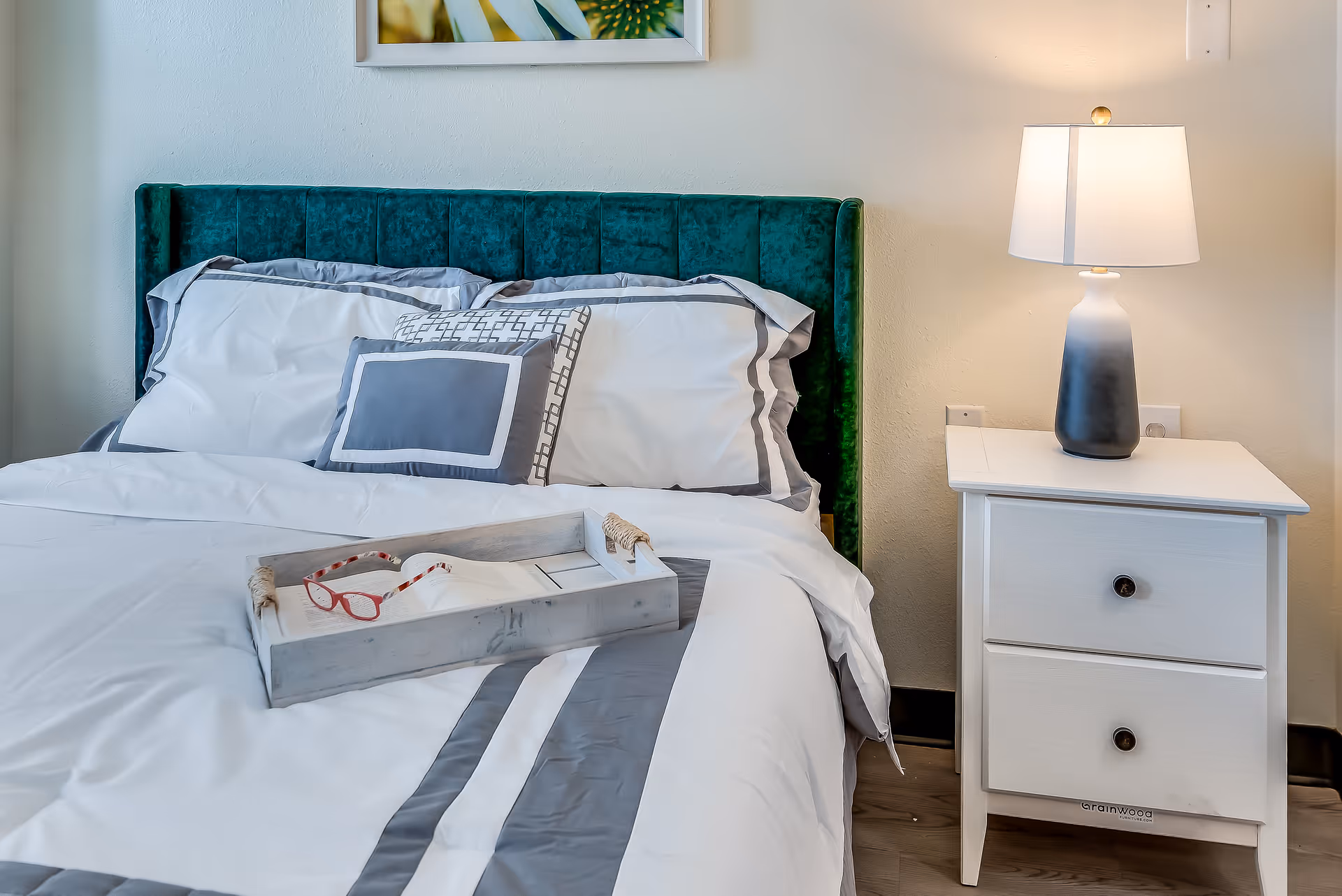 A neatly made bed with white and gray bedding and a green upholstered headboard. On the bed is a wooden tray holding a pair of red reading glasses and an open book. Next to the bed is a white nightstand with two drawers and a modern table lamp with a white shade and a gradient black and white base. A framed floral picture hangs on the wall above the bed.