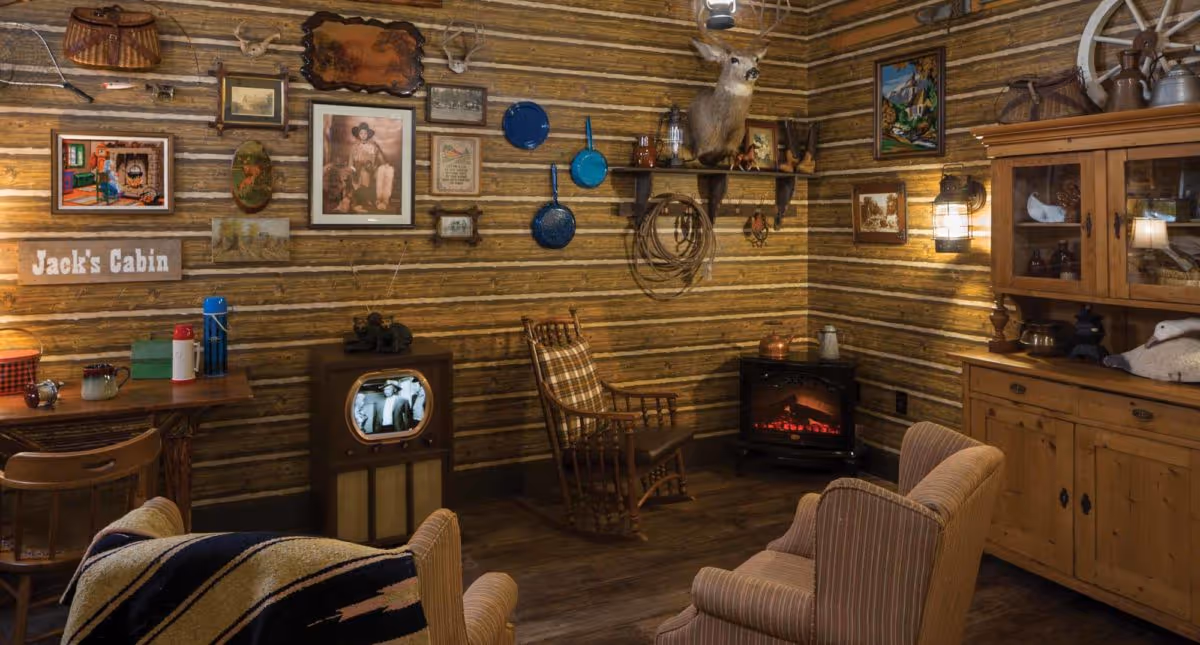 A cozy rustic living room with log cabin style walls decorated with vintage pictures, a mounted deer head, and blue pans. The room features a wooden rocking chair, a small vintage television showing a black and white image, an electric fireplace, and wooden furniture including a cabinet and chairs with striped upholstery. A sign on the wall reads 'Jack's Cabin'.