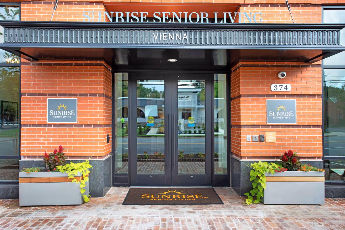 Entrance to Sunrise Senior Living facility in Vienna with double glass doors, brick walls, two planters with green and red plants, and signage displaying the facility name.