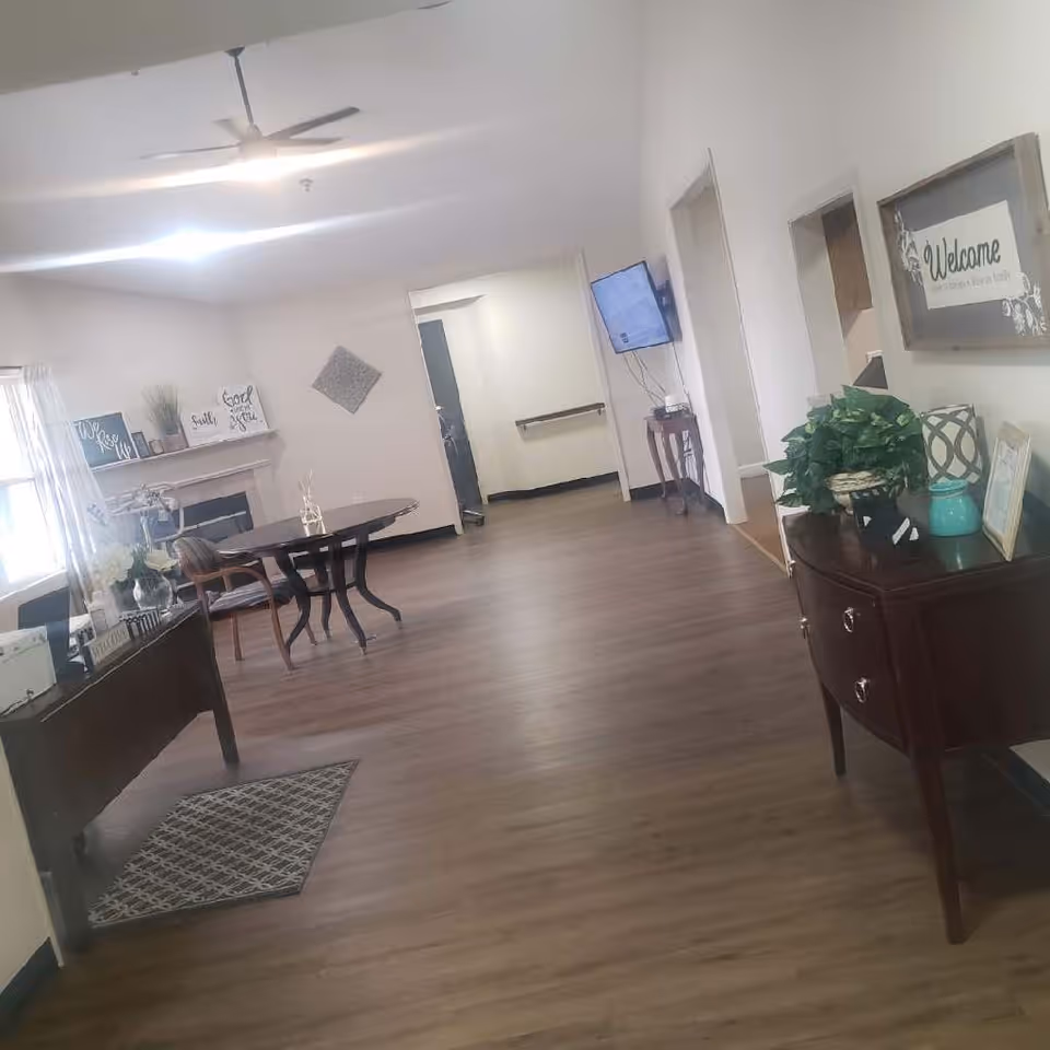 Interior view of a common area in an assisted living facility with wooden flooring, a ceiling fan, a round table with chairs, a wall-mounted TV, decorative plants, and framed wall art including a 'Welcome' sign.