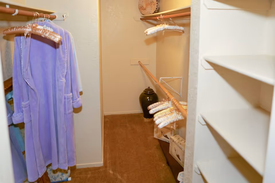 Interior view of a walk-in closet with beige walls and carpeted floor. The closet has wooden hanging rods with several padded hangers and a purple bathrobe hanging on the left side. There are white shelves on the right side and a decorative dark vase placed on the floor at the back of the closet.