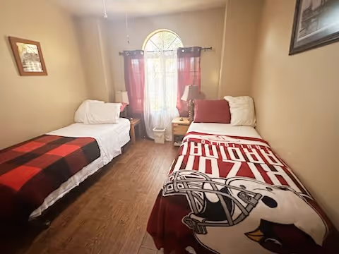 A small bedroom with two single beds placed parallel to each other against opposite walls. Each bed has white pillows and blankets, with one bed covered by a red and black checkered blanket and the other by a red blanket featuring a sports team logo. Between the beds is a window with an arched top, covered by sheer white curtains and red drapes. There are two bedside tables with lamps on each side of the window. The room has wooden flooring and beige walls with a framed picture on the right wall.