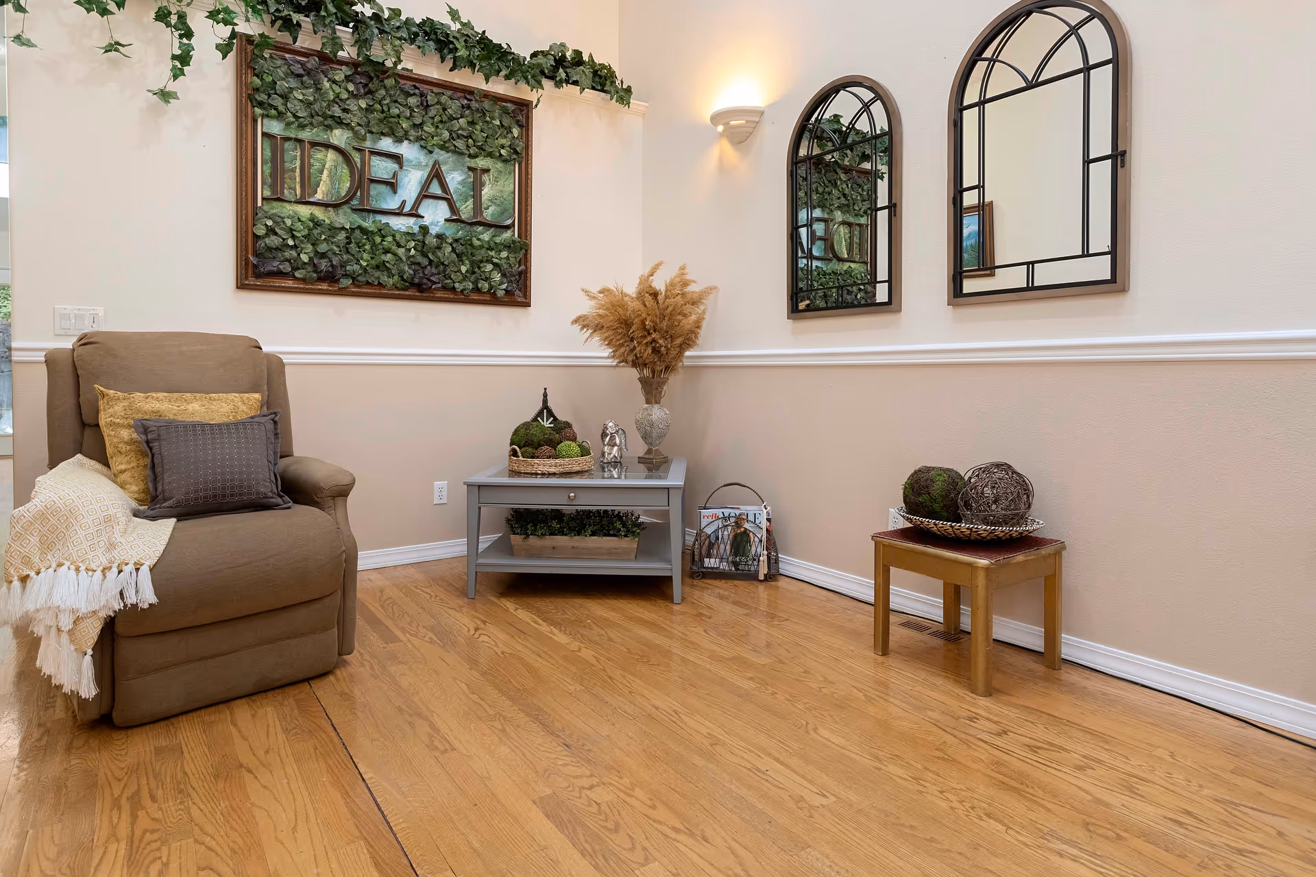 Cozy seating area with an armchair, small tables, decorative mirrors and a framed plant sign reading "IDEAL" on a hardwood floor.