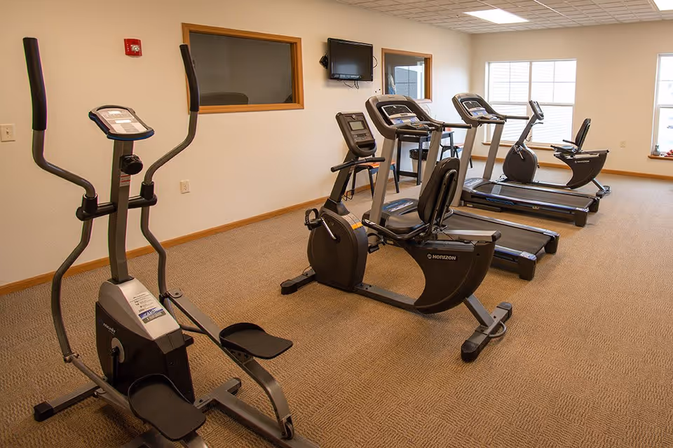 A fitness room with exercise equipment including an elliptical machine, a recumbent bike, two treadmills, and a stationary bike. The room has beige carpet, white walls, two windows, and a wall-mounted TV.