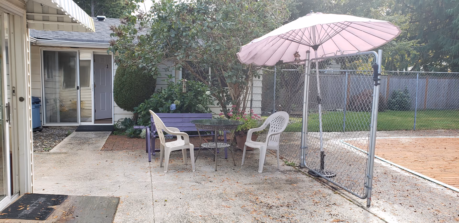 Outdoor patio area with a round glass table, two white plastic chairs, a purple bench, and a pink umbrella attached to a chain-link fence. There is a concrete ground and a building entrance with a sliding glass door and a gray door labeled number 3. Green bushes and trees are visible in the background.