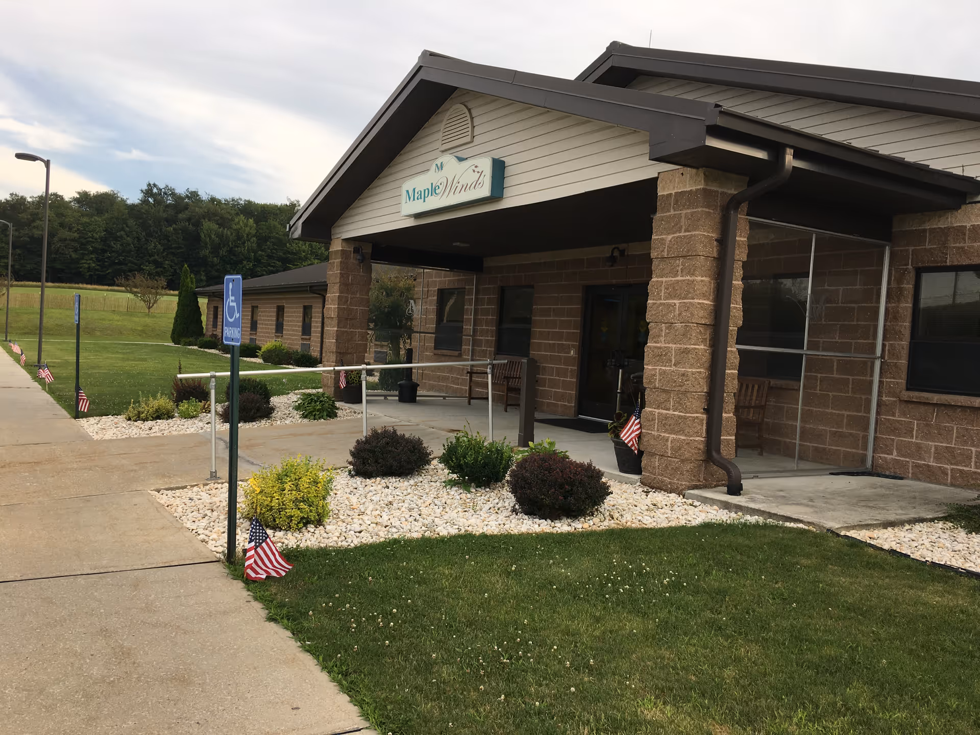 Exterior view of Maple Winds Healthcare And Rehabilitation Center showing the entrance with a covered porch supported by brick columns. There is a wheelchair accessible ramp leading to the door, small landscaped areas with bushes and white rocks, and several small American flags placed around the garden and walkway. The building is surrounded by green grass and trees in the background under a partly cloudy sky.