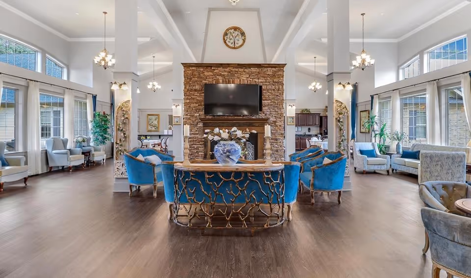 Bright, spacious common lounge with blue upholstered seating arranged around a central stone fireplace and TV, with large windows along the sides.