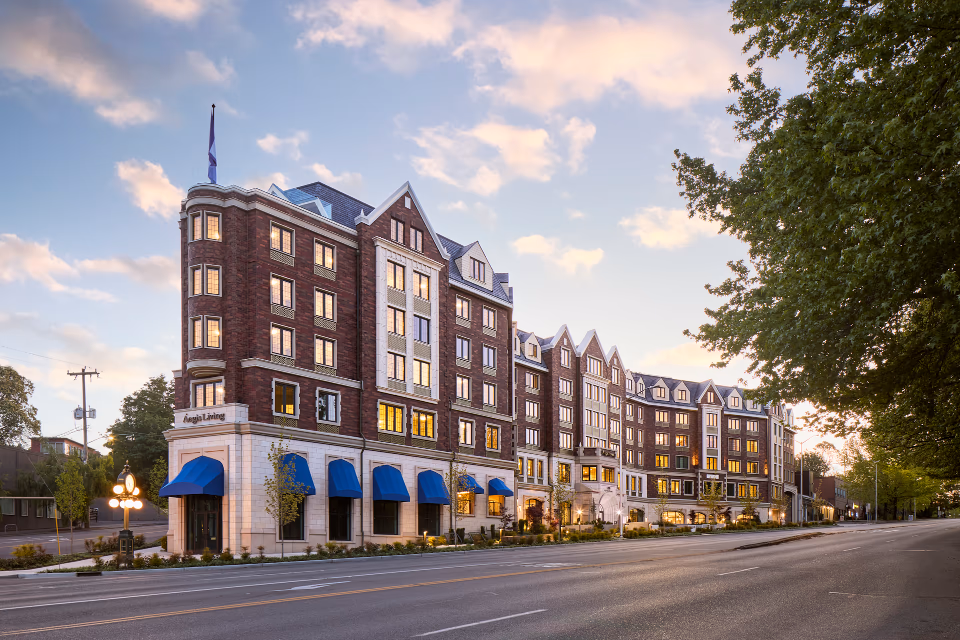 Exterior view of a large multi-story brick building with blue awnings and multiple windows lit from inside during sunset, identified as Aegis Living Laurelhurst, with a tree-lined street in the foreground and a partly cloudy sky.
