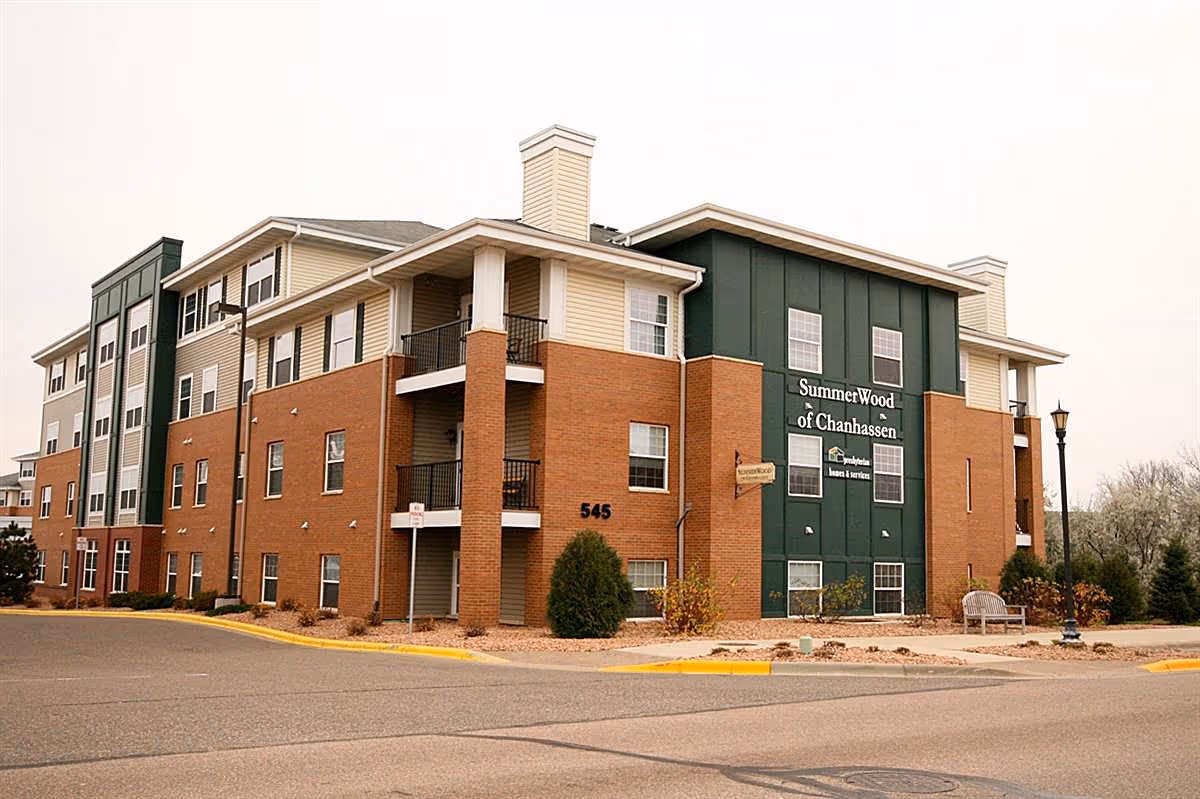 Exterior view of a multi-story senior living facility building named SummerWood of Chanhassen with a combination of brick and green paneling, balconies, and surrounding landscaping.