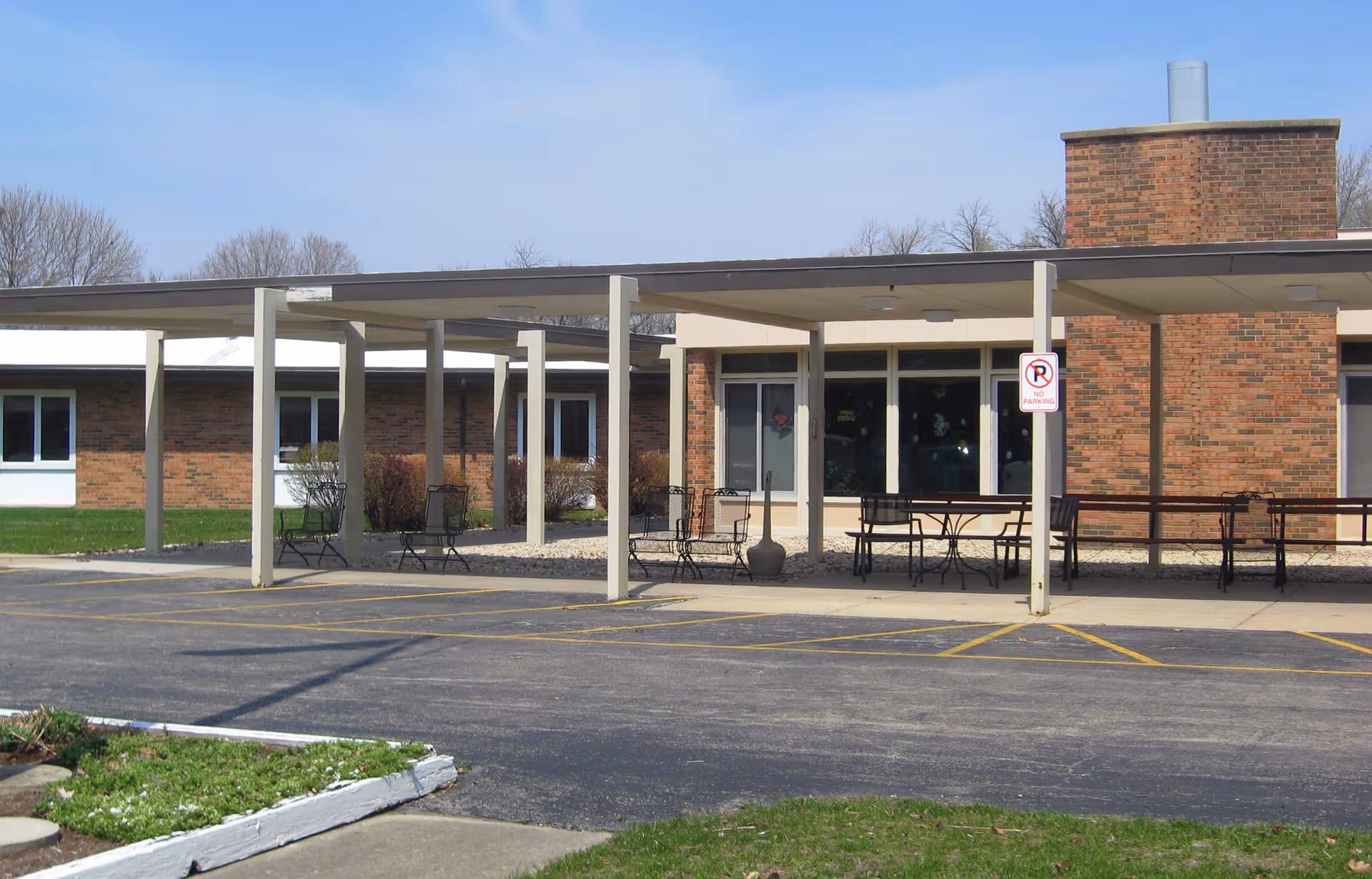 Exterior view of a single-story brick building with a covered walkway supported by white pillars. There are several metal chairs and tables under the covered area. The building has large windows and a chimney. A 'No Parking' sign is visible on one of the pillars. The sky is clear and blue.