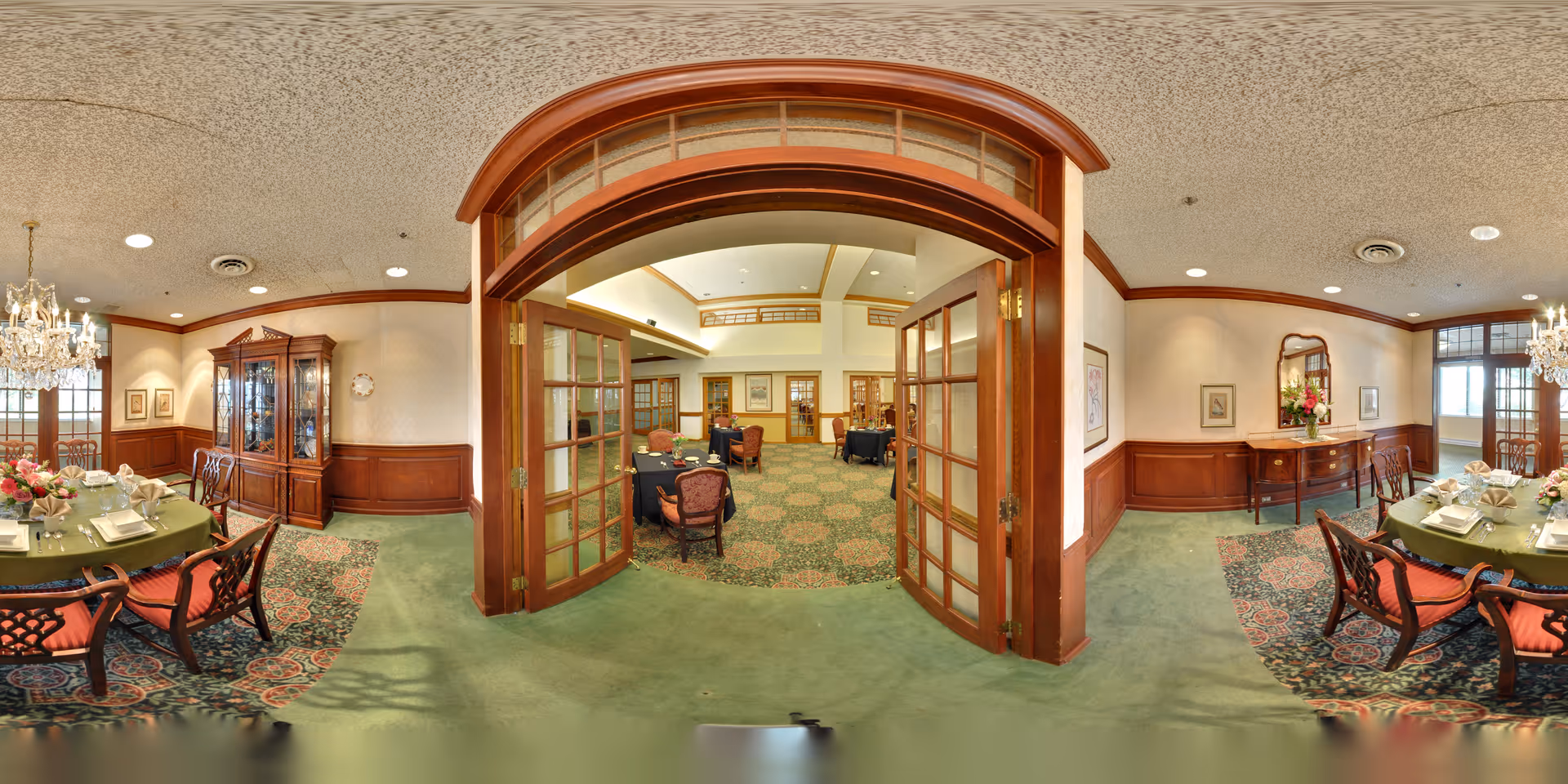 A spacious and elegant dining room with green carpet and wooden paneling. The room features round dining tables set with plates, napkins, and floral centerpieces. There are chandeliers hanging from the ceiling, a wooden china cabinet, and a sideboard with a mirror and flowers. French doors open into another room with additional seating and tables.