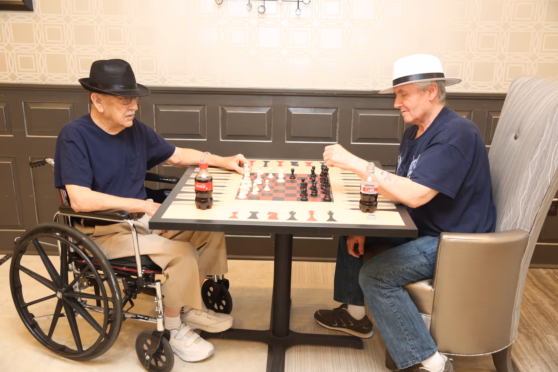 Two elderly men wearing hats are playing chess at a table in an indoor setting. One man is seated in a wheelchair, and the other is sitting in a cushioned chair. Both have bottles of Coca-Cola on the table. The background features a patterned wall with dark paneling.