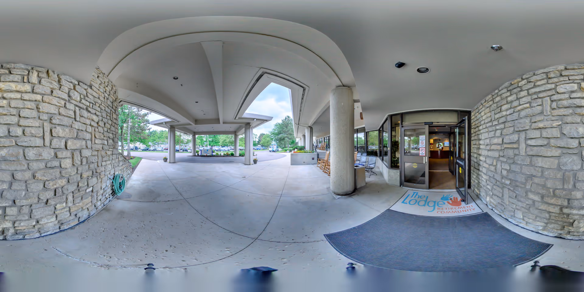 Covered entrance area of The Lodge Retirement Community with stone walls, concrete floor, and large pillars. There is a welcome mat with the facility's name and logo near the glass entrance doors. Outside, trees and parked cars are visible.