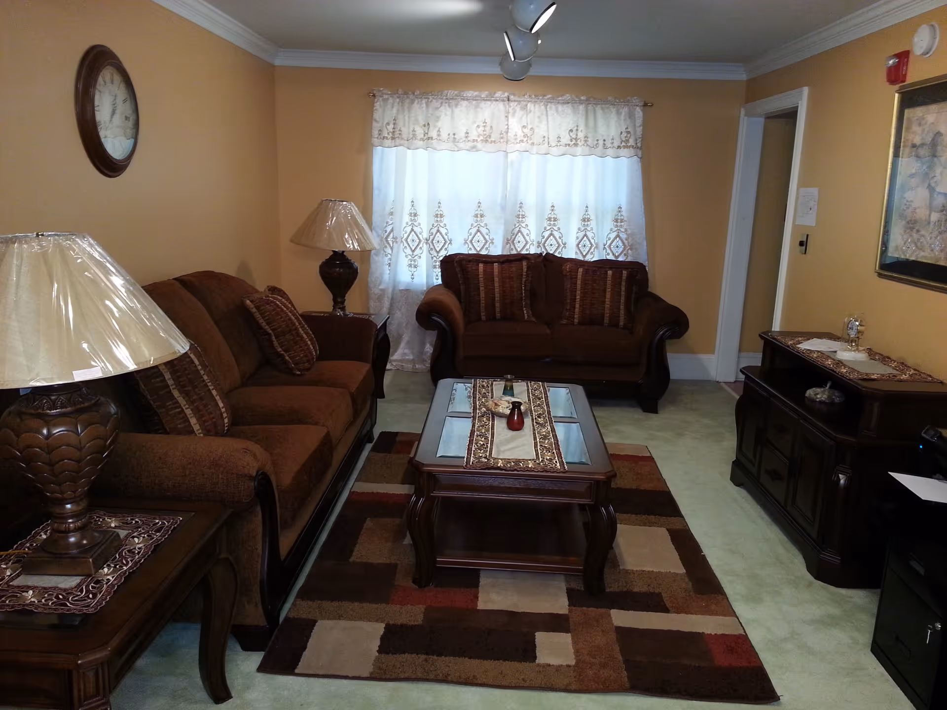 A cozy living room with two brown upholstered sofas, a wooden coffee table with decorative runners and small vases, two matching table lamps with beige lampshades, a patterned area rug, a wall clock, and a framed painting on the wall. The room has beige walls and a window with white lace curtains.