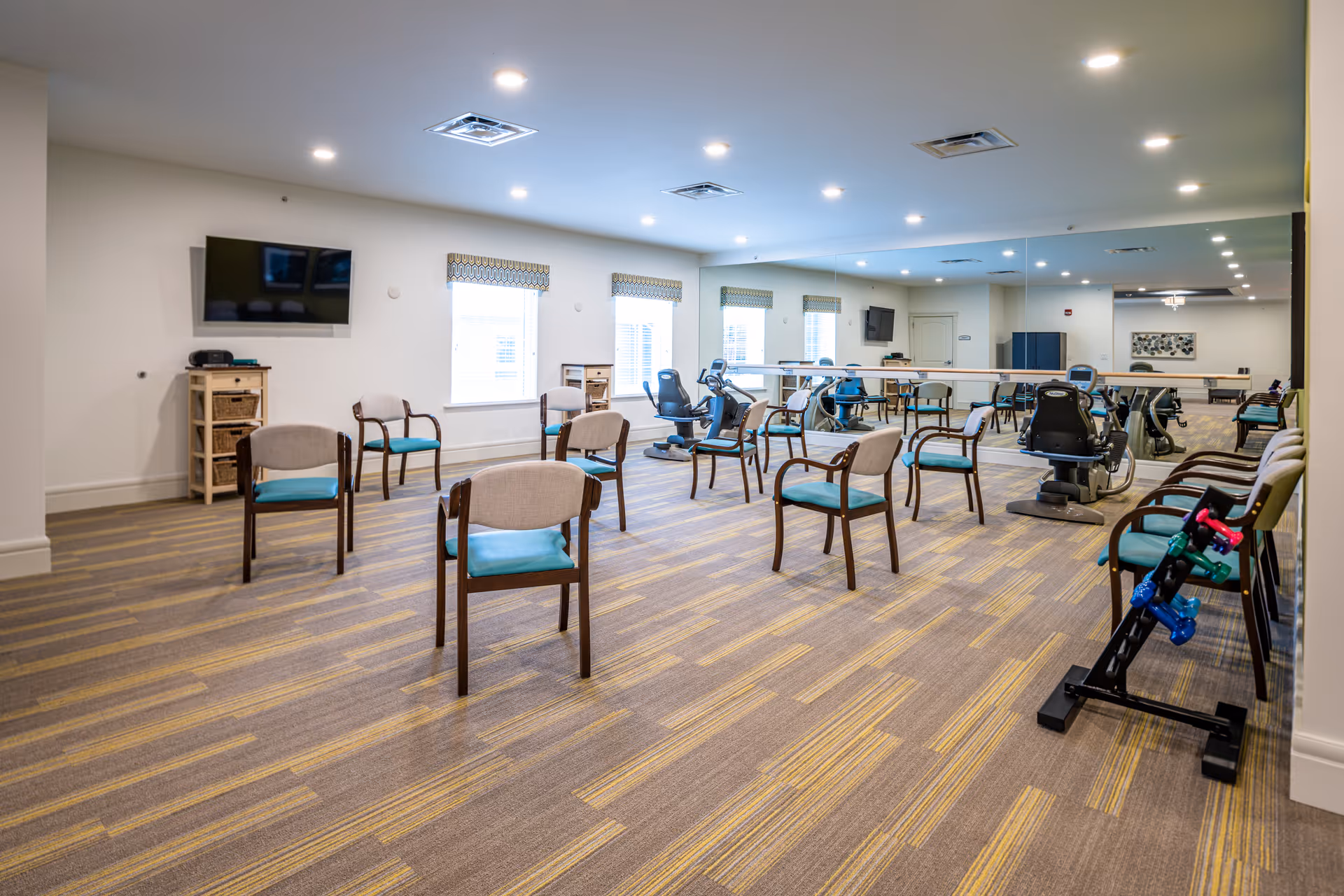 A spacious exercise and activity room with multiple chairs arranged in a circle, exercise equipment including stationary bikes, a large wall mirror, a wall-mounted TV, and windows with patterned valances allowing natural light to enter.
