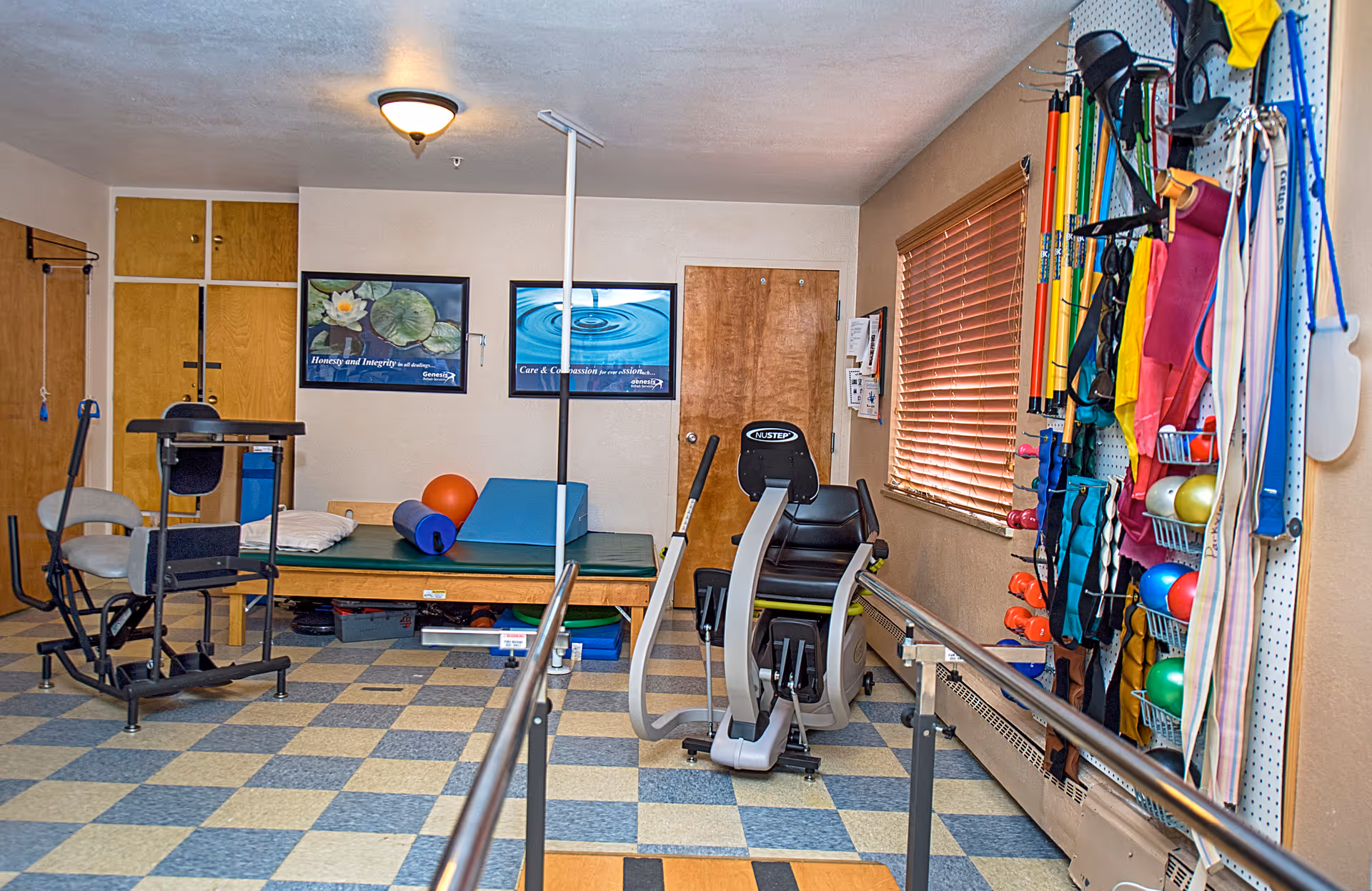 A therapy or exercise room in a senior care facility with various rehabilitation equipment including a recumbent exercise bike, parallel bars, therapy mats, exercise balls, resistance bands, and other physical therapy tools hanging on the wall. The room has checkered flooring, beige walls, a window with wooden blinds, and two framed motivational posters on the wall.