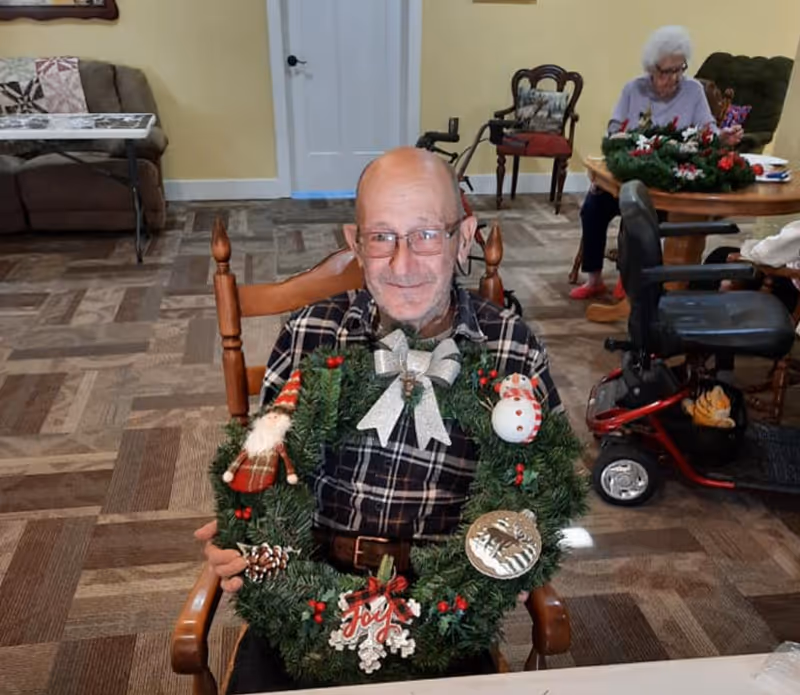 An elderly man wearing glasses and a plaid shirt is sitting in a wooden chair indoors, holding a decorated Christmas wreath with ornaments including a snowman, a Santa figure, and a silver bow. In the background, an elderly woman is seated at a round table with a similar wreath and other items. The room has patterned carpet flooring, yellow walls, and various chairs and furniture.