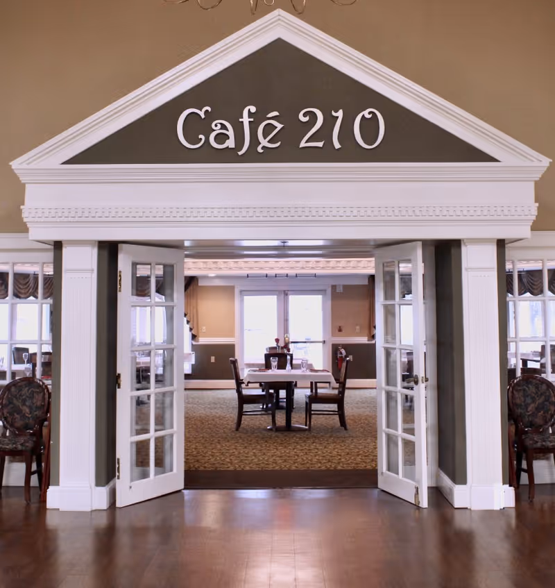 Entrance to Café 210 with double glass doors open, showing a dining area with tables and chairs inside a senior living facility.