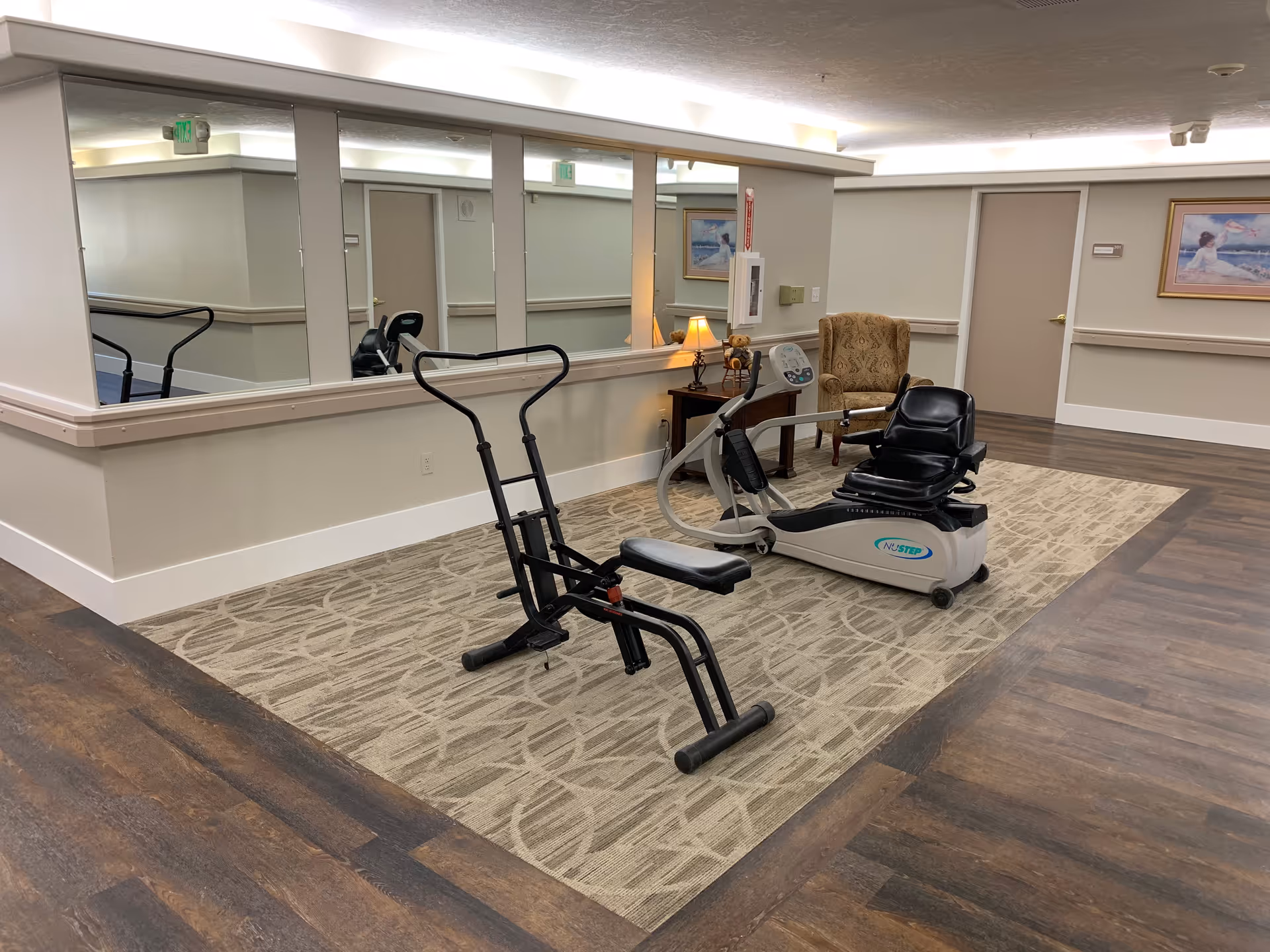 A small exercise area inside Ridgeview Gardens Assisted Living featuring two exercise machines on a patterned carpet. The area is surrounded by wood flooring and has large mirrors on one wall. There is a cushioned armchair and a small table with a lamp and teddy bears next to the exercise machines. The walls are light-colored with framed artwork and a closed door in the background.