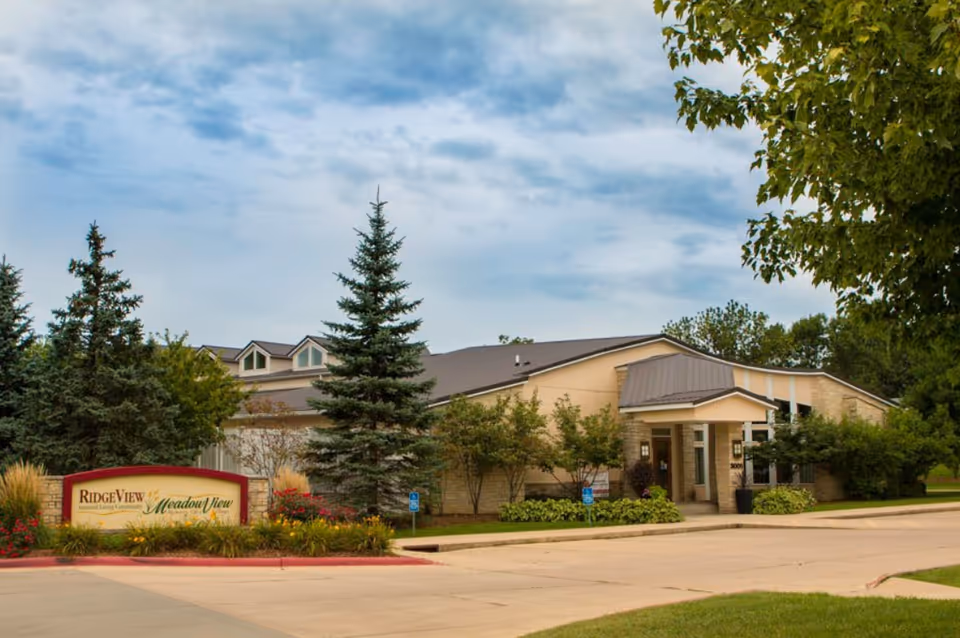 Front exterior of a single-story senior living building with trees, landscaping, and a stone sign by the driveway.