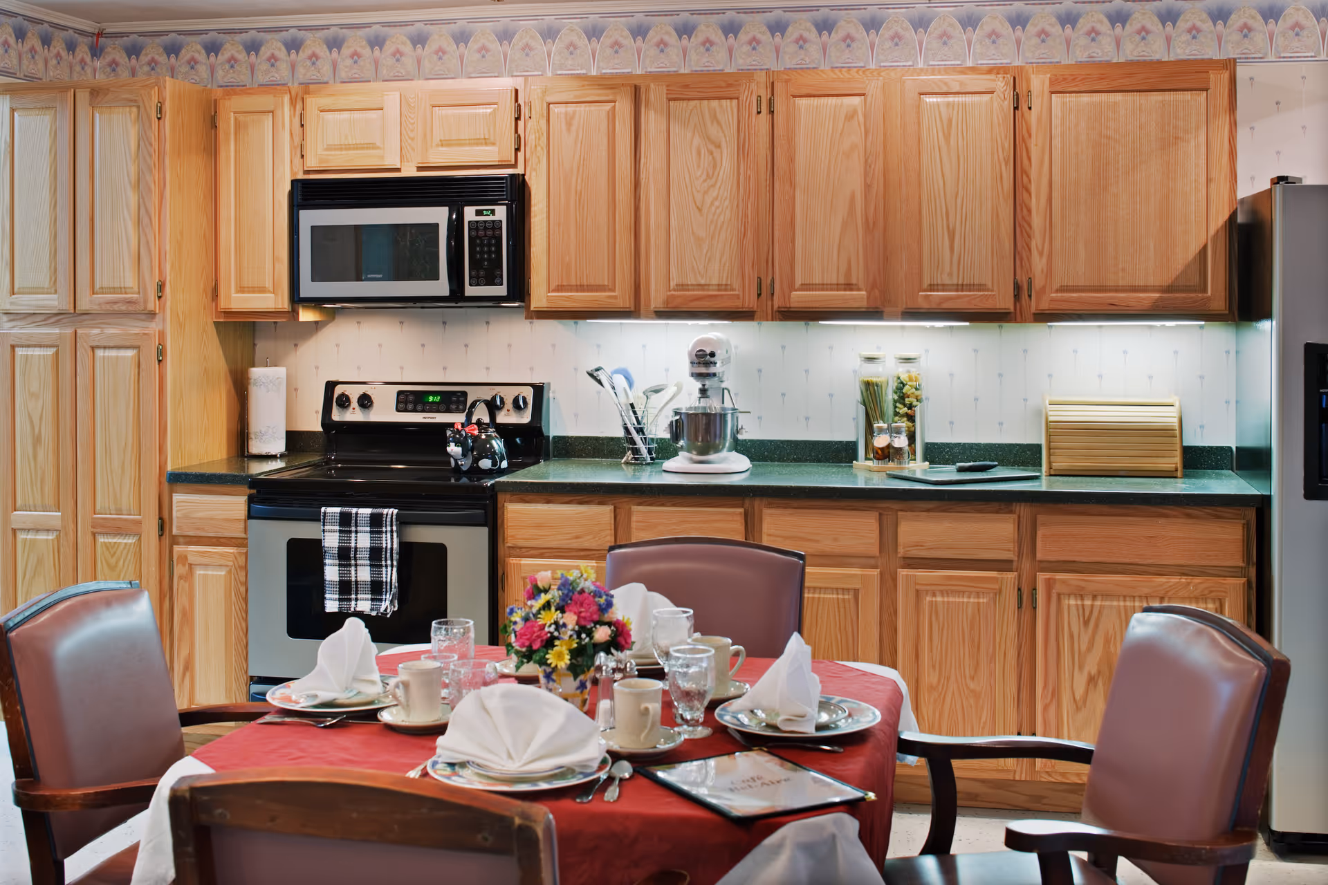 A kitchen area with wooden cabinets, a microwave, stove, and refrigerator. In front of the kitchen counter is a dining table set with a red tablecloth, white napkins, cups, glasses, and a floral centerpiece. Four chairs surround the table.