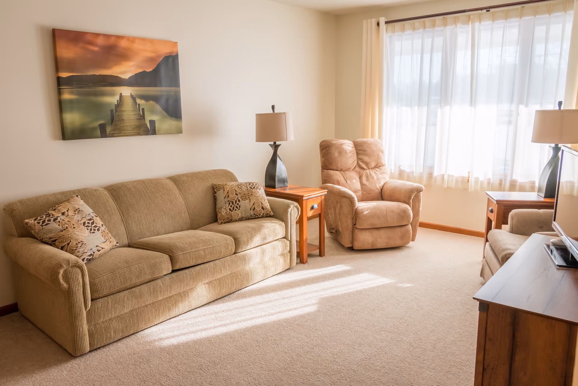 A cozy living room with a beige sofa, a matching armchair, two wooden side tables with lamps, a large window with sheer curtains letting in natural light, and a wall painting of a wooden pier extending into a calm lake at sunset.